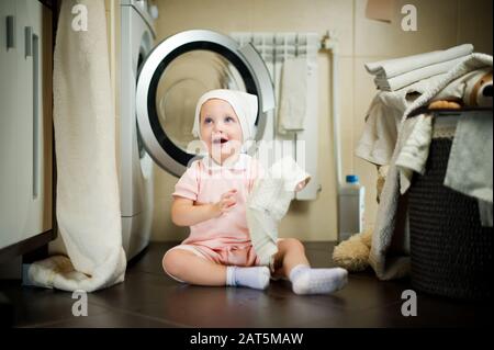 baby girl sitting on the floor near the washing machine in the bathroom with a towel in her hand Stock Photo