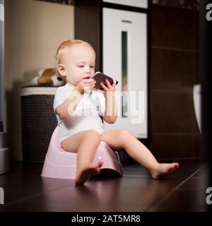 child is holding a smartphone in her hands sitting on a pot in the bathroom. Toned Stock Photo