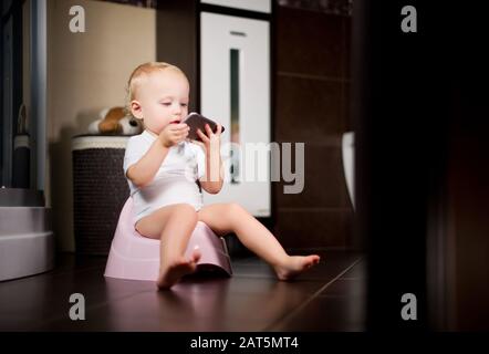 child is holding a smartphone in her hands sitting on a pot in the bathroom. Toned Stock Photo