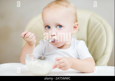 baby girl inquiringly looks at the camera, holding a spoon and about to eat porridge Stock Photo