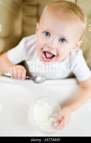 A little blue-eyed girl in a feeding chair places a spoon with rice porridge in her mouth. View from above, vertical photo Stock Photo