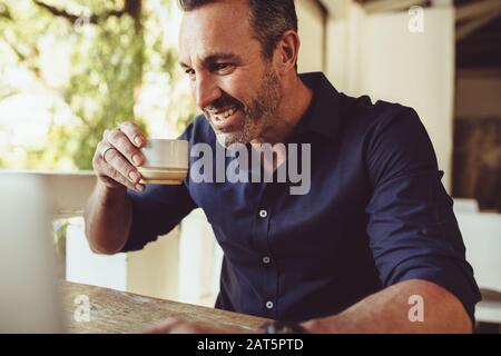 Smiling man sitting by the table in cafe with laptop drinking coffee. Man having a cup of coffee and looking at his laptop computer. Stock Photo