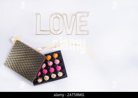 Top view festive composition of black and golden box with allsorts chocolate sweets with multicolored cream filling and wire word Love on white backgr Stock Photo