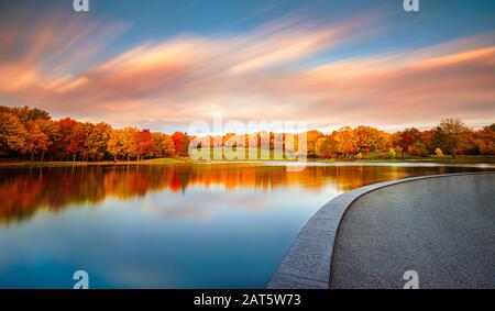 The beautiful orange foliage of autumn and the blue sky with streaking, colorful clouds are reflected into the perfectly still surface of the lake. Stock Photo