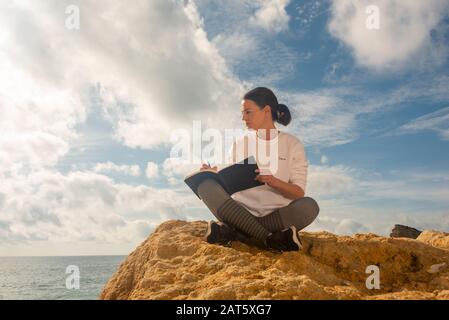Woman hand writing on a notepad with a pen, sitting cross legged outside on a rock by the sea. Stock Photo