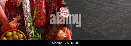 top view of delicious meat platter served with olives and herbs on wooden black table, panoramic shot Stock Photo