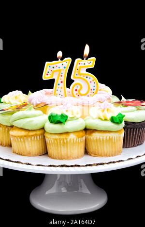 Birthday cake with pastel colored butter cream icing surrounded by cupcakes. A number 75 candle is burning in the middle of the cake. Stock Photo