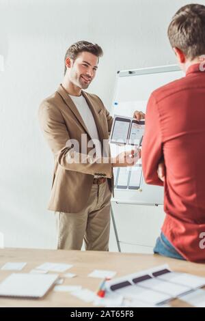 Selective focus of smiling designer showing screens of mobile responsive website development to colleague in office Stock Photo