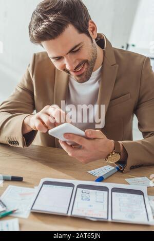 Selective focus of smiling designer using smartphone beside wireframe sketches of mobile website on table Stock Photo