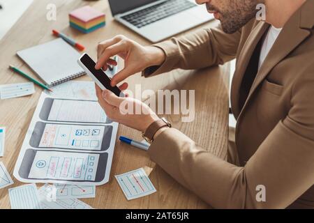 Cropped view of designer using smartphone with mobile web prototype on table Stock Photo