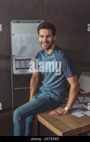 Handsome ux designer smiling at camera while sitting on table with sketches Stock Photo