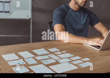 Cropped view of ux developer using laptop beside designer sketches on table, selective focus Stock Photo