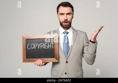 displeased businessman looking at camera while holding chalkboard with copyright inscription isolated on grey Stock Photo