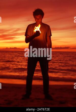 A young man stands on a beach at sunset and looks at his burning smart phone as flames illuminate his face Stock Photo
