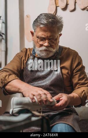 senior, bearded cobbler in leather apron repairing shoe in workshop Stock Photo