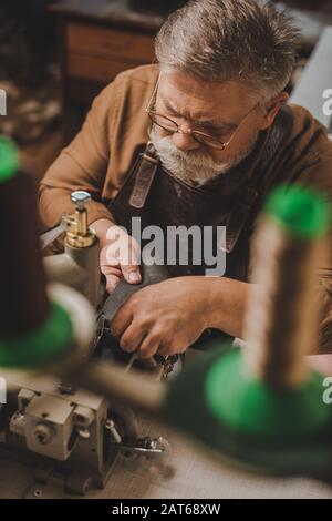 selective focus of senior shoemaker sewing leather on sewing machine in workshop Stock Photo