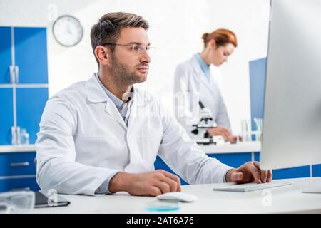 selective focus of molecular nutritionist using computer and his colleague using microscope on background Stock Photo