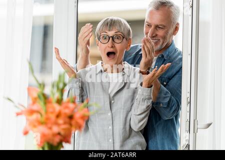 selective focus of smiling man and shocked woman looking at bouquet in new house Stock Photo