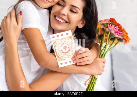 happy woman hugging daughter holding flowers and mothers day card with i love you mommy inscription Stock Photo