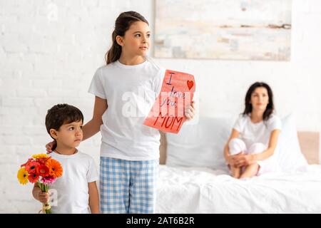 cute brother and sister holding i love you mom card and flowers, while mother sitting in bed Stock Photo