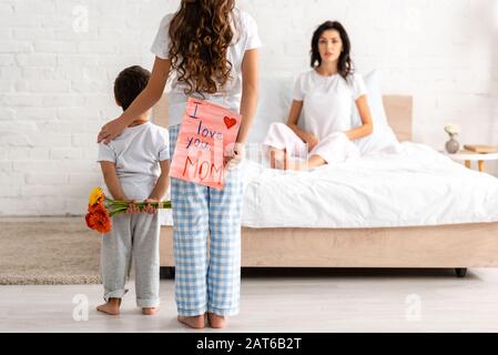 back view of brother and sister holding flowers and i love you mom card, and surprised woman sitting in bed Stock Photo