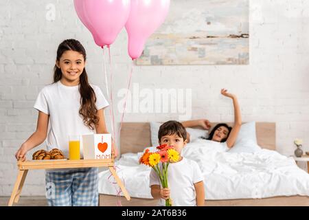 cute girl holding tray with breakfast and mothers day card with heart sign and mom lettering, and boy holding bouquet while mother stretching in bed Stock Photo