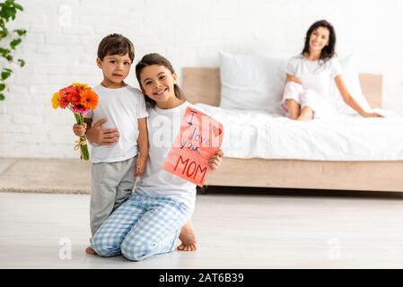 selective focus of children holding i love you mom card near mother sitting in bed Stock Photo