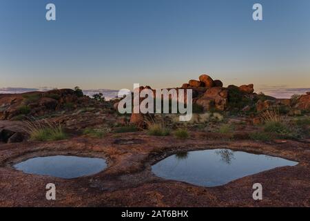 Two desert rock pools in the foreground with rock formations in the background framed in a beautiful painted sunset at the Devil's Marble, Australia. Stock Photo