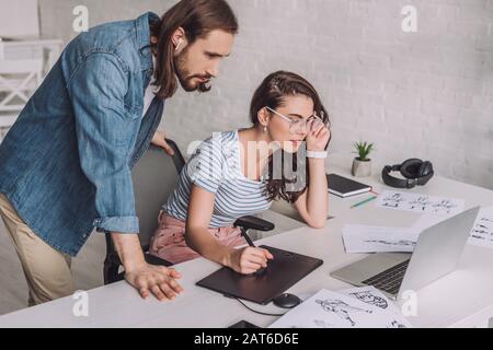 illustrator looking at laptop near coworker in glasses and sketches Stock Photo