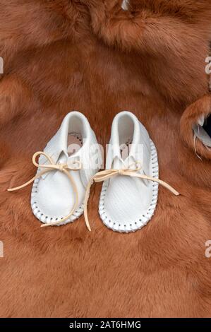 A pair of white leather baby moccasins are displayed on a brownish red rabbit pelt with bokeh effect. Stock Photo