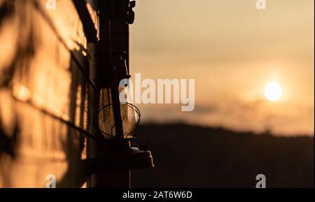 Old oil lamp hanging outside on a wall of a wooden hut in the evening while sunset Stock Photo