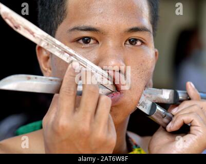 Handsome young Thai Chinese Taoist devotee (mah song) pierces his left cheek with two knives during Phuket's Nine Emperor Gods Festival. Stock Photo