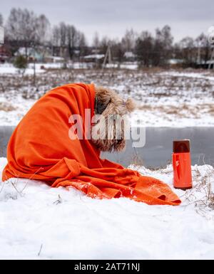 A dog in a bright orange plaid sits on the Bank of the river, facing the camera. Next to it is an orange thermos. Stock Photo