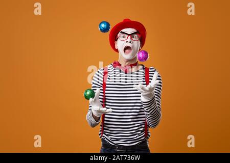 Mime in vest and red hat juggles with colorful balls on orange empty background Stock Photo