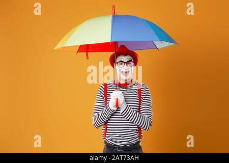 Funny mime with multi-colored umbrella on empty orange background in studio Stock Photo