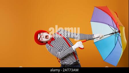 Mime man with multi-colored umbrella during wind isolated on empty orange background in studio. Stock Photo