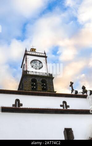 The outdoor facade of St. Sebastian Church, Igreja Matriz de Sao Sebastiao, in Ponta Delgada, Azores, Portugal. White clock tower from below with blue sky and clouds above. Sunset clouds. Stock Photo