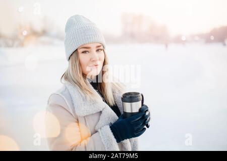 Portrait beautiful young girl in warm clothes and hat in winter holds thermos mug with hot tea, bokeh blurred background Stock Photo