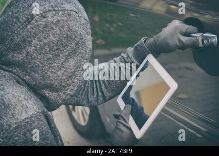 Hooded thief tries to break the car's security systems with tablet. Hacking modern car concept Stock Photo