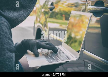 Hooded thief tries to break the car's security systems with laptop. Hacking modern car concept Stock Photo