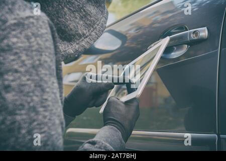 Hooded thief tries to break the car's security systems with tablet. Hacking modern car concept Stock Photo