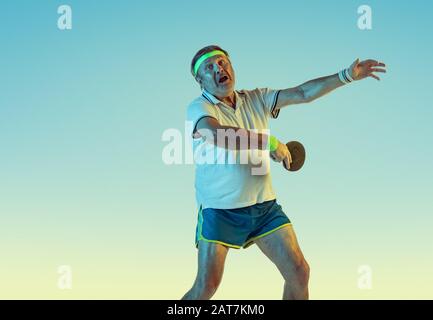 Senior man playing table tennis on gradient background in neon light. Caucasian male model in great shape stays active, sportive. Concept of sport, activity, movement, wellbeing, healthy lifestyle. Stock Photo