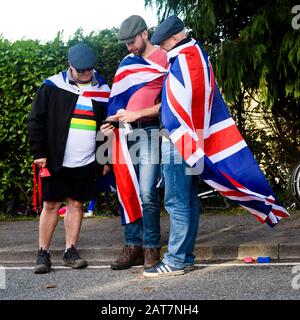 3 men looking at phone, standing at side of road, wearing flat caps & Union Jack flags, spectators at sports event - Harrogate, Yorkshire, England UK Stock Photo