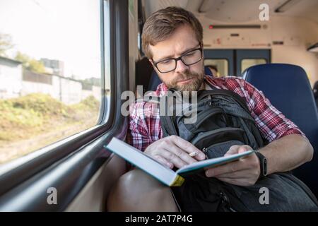 Focused bearded man riding in a train, reading a book and making notes with a pencil. Stock Photo