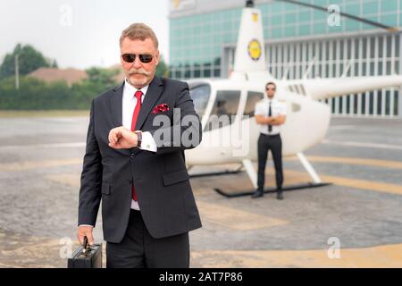 Elderly Businessman manager with Briefcase watching watch to checking time when he arrive destination at helipad in airport with helicopter and captai Stock Photo