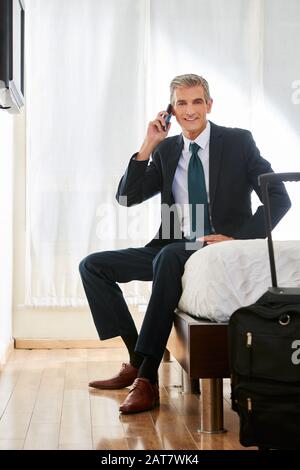 Smiling businessman in hotel talking on the smartphone with his smartphone on the bed Stock Photo