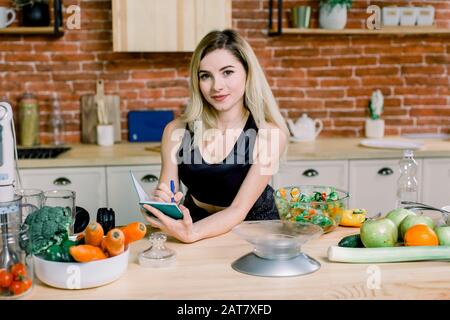 Young pretty woman in the kitchen at home writing shopping list and watching at camera. Modern kitchen, fresh vegetables and healthy food on the Stock Photo