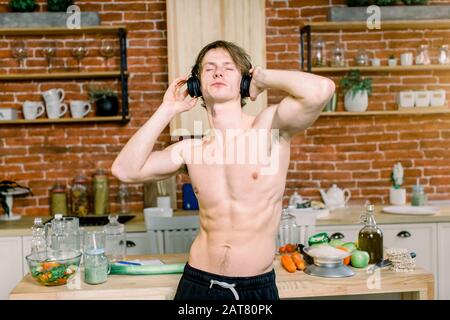 Handsome man drinking green smoothie in kitchen. Young man is listening to music in earphones, drinks smoothie while dancing. Healthy lifestyle and Stock Photo