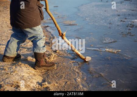 Close-up of legs Small four years caucasian boy standing near riverside and playing with long wooden stick in sunny winter day Stock Photo