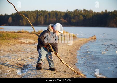 Small four years caucasian boy standing near riverside and playing with long wooden stick in sunny winter day Stock Photo